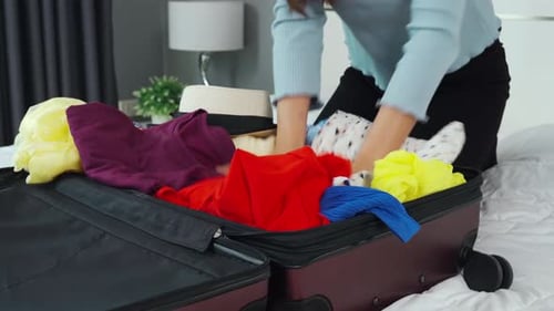 Young Adult Packing Suitcase with Colorful Clothes on Bed