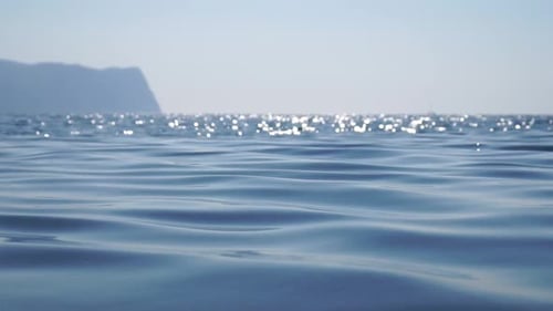 Sea Water Surface Camera Flies Over the Calm Azure Sea with Volcanic Rocky Shores on Background