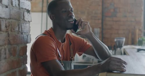 Cheerful African American Man Barista in Apron Speaking to Client on Mobile Phone in Modern Cafe