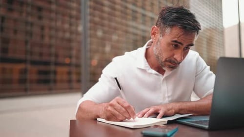 Mature businessman with beard, user on laptop pc computer and make notes sit at cafe outdoors