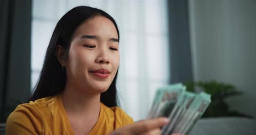 Happy young Asian woman smiling confidently counting money at home.