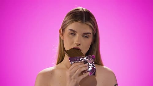 Woman Eats Chocolate Bar Against Pink Backdrop