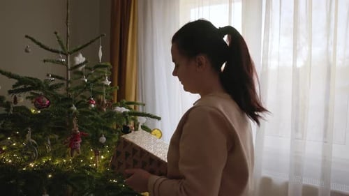 A Girl, Holding a Gift, Examines the Decorations Hanging on the Christmas Tree - Medium Close Up