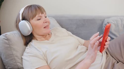 Elderly Woman Enjoying Time at Home with Headphones Smiling While Using Smartphone on a Cozy Gray