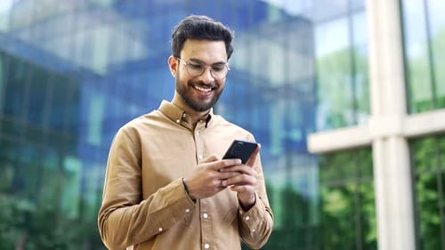 Confident businessman standing near modern glass business office building holding smartphone in hand