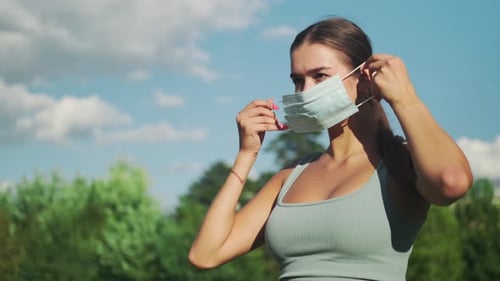 A beautiful girl in sportswear in a sunny city park puts a medical mask on her face. Side view