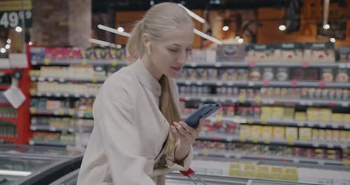 Young Lady Shopping in Supermarket Using Smartphone App and Looking at Food in Trolley