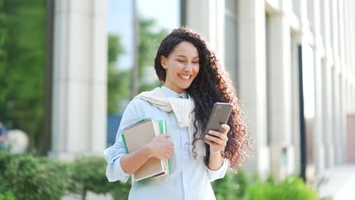 Smiling female student with books in hands using smartphone standing in campus space near university