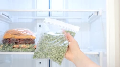 Female hand holding zip bag with frozen green peas against open freezer background.