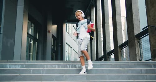 Cheerful Caucasian schoolboy in headphones dancing walking with backpack after classes. Portrait
