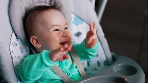 Funny kid sitting in a baby chair is fed from spoon. Childs waves hands and smiles. Close up.