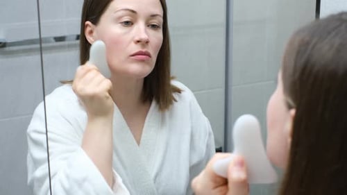 Woman Massaging Face with Gua Sha in Bathroom Mirror