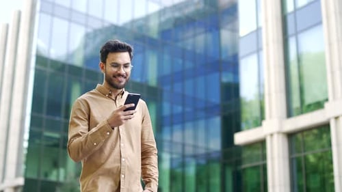 Confident businessman walking near modern glass business office building holding smartphone in hands