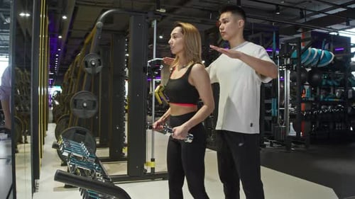 Young Asian Gym Instructor is Helping an Attractive Athlete Do Some Hand Lifting Exercises Using