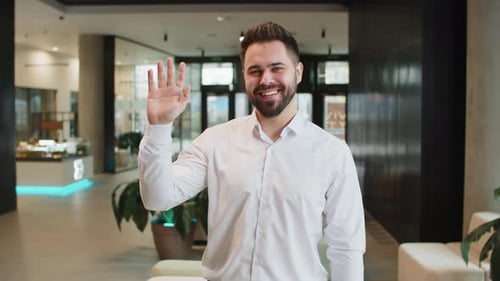 Happy Young Caucasian Businessman Looking at Camera and Waves Hand Hello in Modern Office Lobby