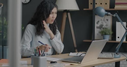 Ambitious Asian Lady Looking at Laptop Screen and Writing Information in Notebook at Desk in Office