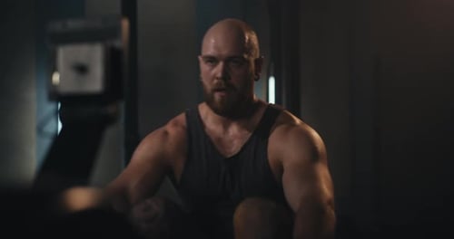Closeup of a Bearded Man in a Tank Top Intensely Working Out on a Rowing Machine in a Modern Gym