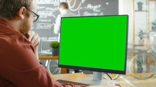 Man Works on the Personal Computer with Green Mock-up Screen. In the background Creative Office wit