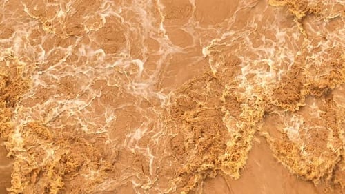 Drohnen-Luftaufnahme von turbulentem, schlammigem Hochwasser, das über Felsen kaskadiert.