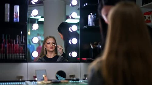 Young blond girl with bright make-up sitting in the chair looking in the mirror with lamps