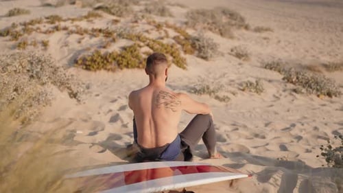 Surfer Contemplates the Waves While Sitting on a Sand Dune with His Surfboard
