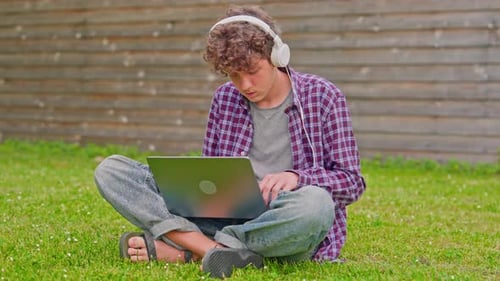 Curly Teenage Boy Listening to Music on Headphones and Works on a Laptop While Sitting in Nature in