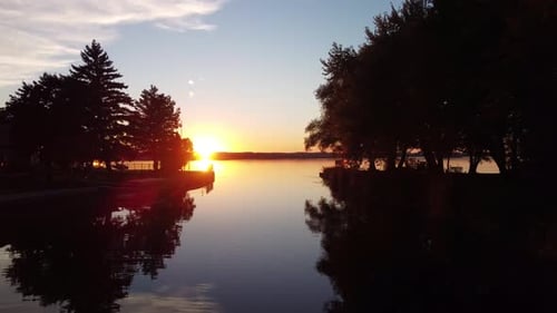 Push in drone view at the sun setting above a lake and trees. Dynamic shot along a channel with park