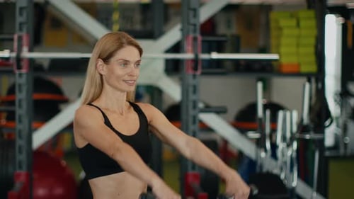 Femme d'âge moyen positive soulevant des haltères faisant de l'exercice au gymnase et souriant au Ralenti dans un espace vide