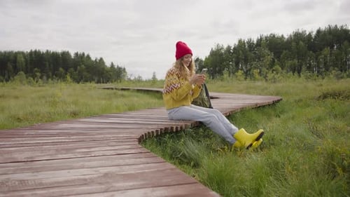 Stylish Girl Sits with a Phone in Her Hands on a Wooden Path While Walking Through the Forest