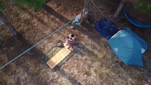 Young woman sitting reading a book close to camping tent in wild pine forest. Aerial top-down descen