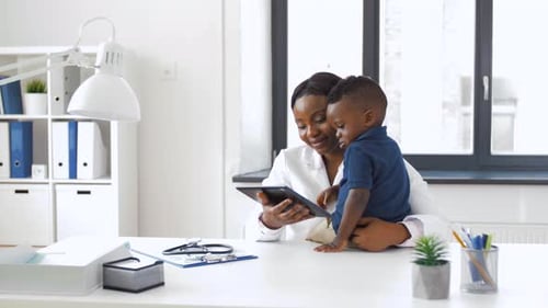 Happy african american doctor showing tablet to baby boy patient at clinic