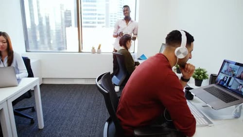 Businessman uses laptop in open plan office for video conference with colleagues