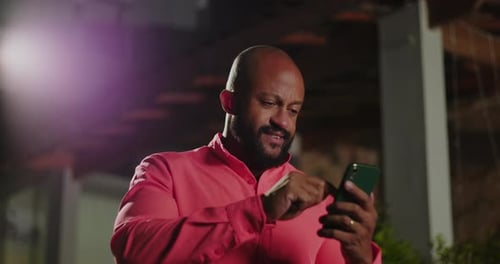 Man of African descent tapping on smart phone and smiling casually outdoors at night, coral shirt