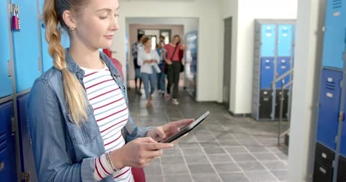 Using tablet, teenage girl standing in high school hallway near lockers