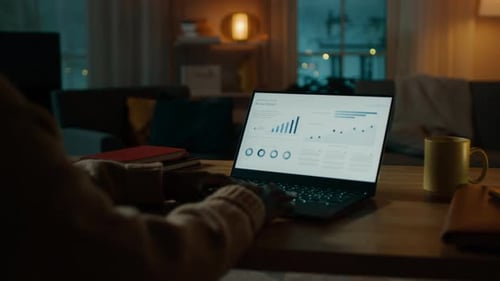 Over the Shoulder Shot: Woman Sitting at Her Desk Works on a Laptop that Shows Statistics and Infog