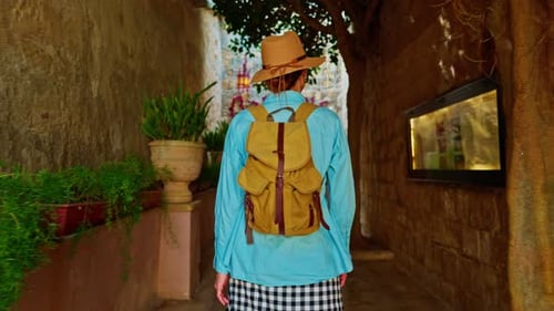 A Female Tourist in a Hat and with a Backpack Walks Along the Traditional Narrow Old Streets in