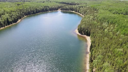 Aerial above a large blue colored lake with waves that is surrounded by a vast green forest.