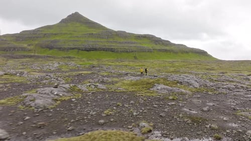 Two hikers traverse rugged terrain in the Faroe Islands under cloudy skies
