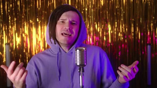 A White Male Hiphop Singer with Dreadlocks in Casual Clothes Reads Rap Into a Microphone Standing on