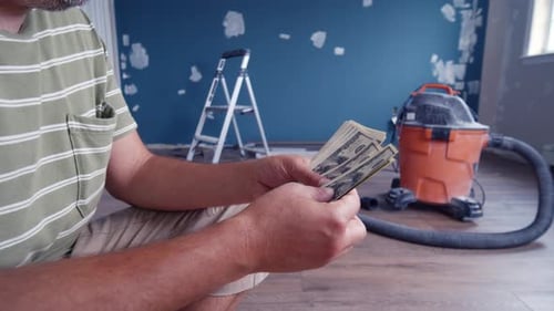 Young Man Counting Money In Renovated Room Surrounded By Tools Painting Supplies