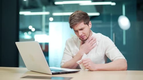 Sick IT programmer blowing running nose sneeze and cough while working on laptop computer at office.