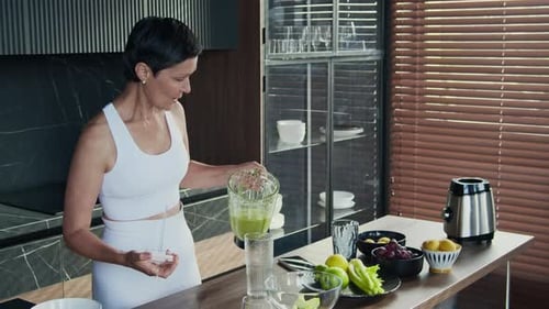 Middle-aged Sporty Woman Humming Song Pouring Green Smoothie at Home