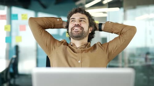 Happy businessman relaxes with hands behind head sitting at workplace in business office. Smiling
