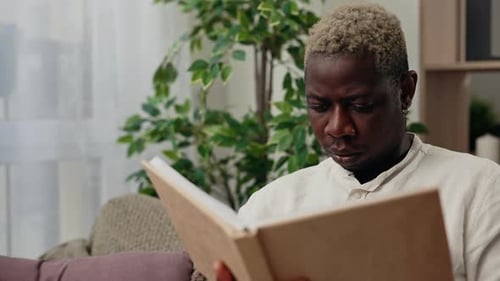 Close Up View of the African American Man Reading a Book After Work Sitting on the Sofa in His