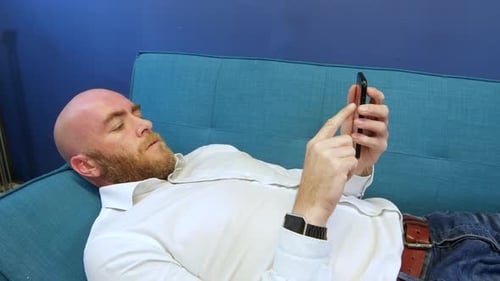 Close shot of man Lying On Sofa And Using Mobile Phone. The bald male has a beard and is wearing a w