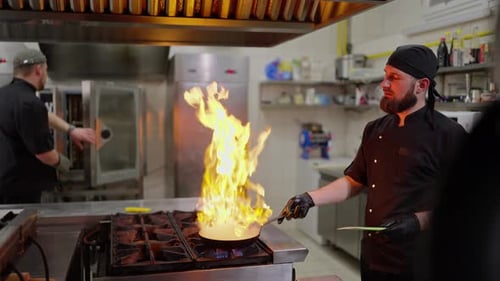 Side View of a Confident Male Chef in a Black Uniform Pouring Alcohol Onto a Hot Frying Pan with a