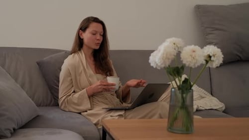 Side View of Young Woman in Bathrobe is Sitting on Sofa Drinking Coffee and Making Video Call Using