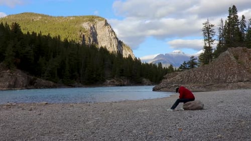 Hiker wearing a red jacket relaxing beside a mountain river.
