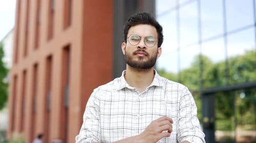 Portrait of a young smiling man standing with crossed arms on the street near an modern office