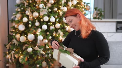 Woman Smiles Holding Christmas Gift Near Decorated Tree